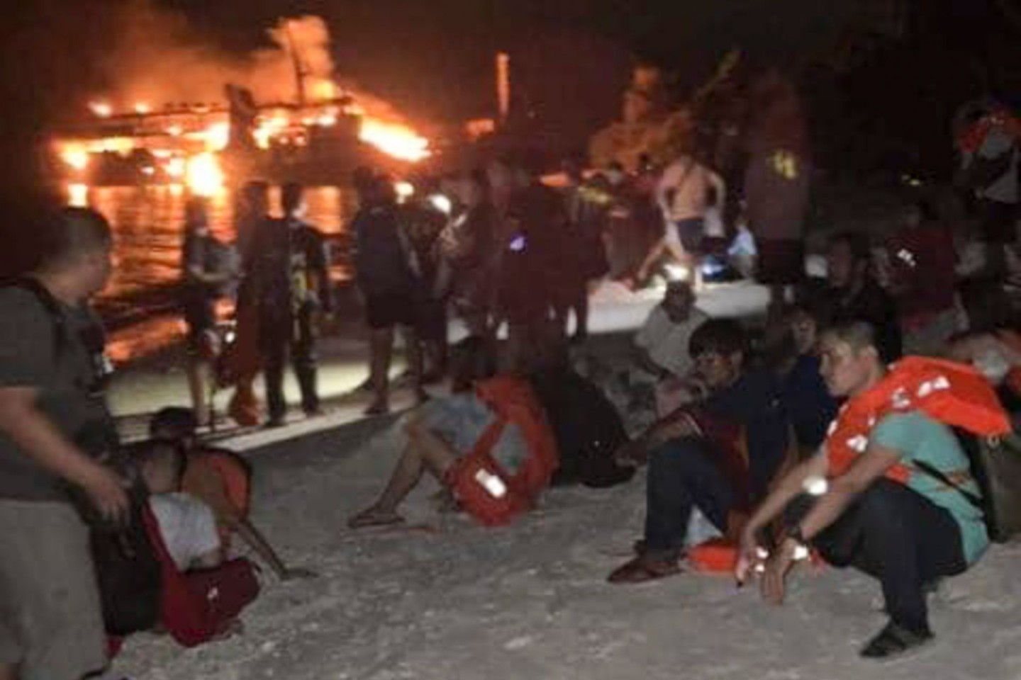 Nach ihrer Rettung ruhen sich Überlebende an Land aus. Im Hintergrund ist die brennende Fähre zu sehen.